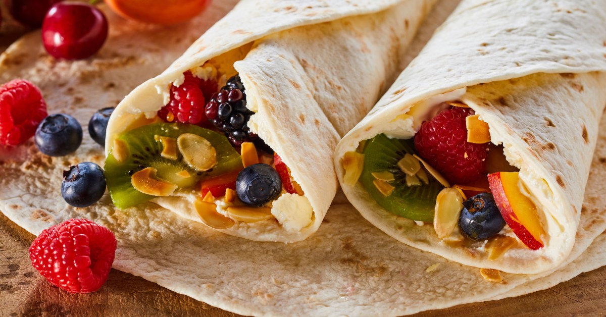 Two tortillas, on top of a larger tortilla, rolled and wrapped around various fruits including strawberries, kiwis, and various berries.