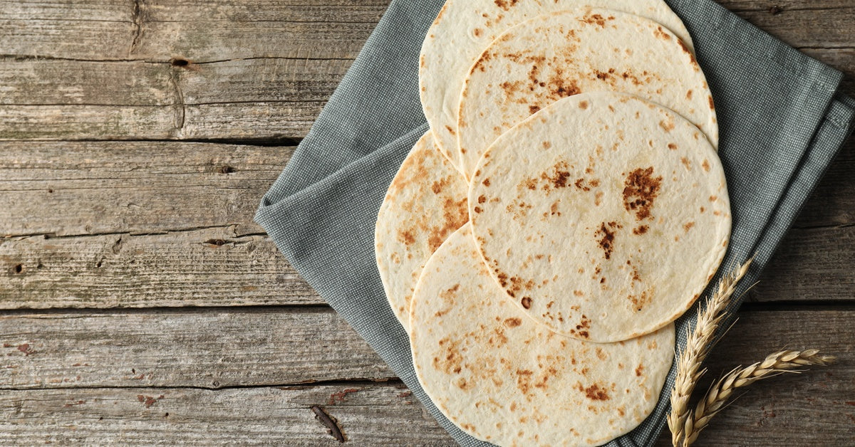 A stack of homemade tortillas sits on a gray cloth napkin next to two stalks of wheat on a rustic wooden table.