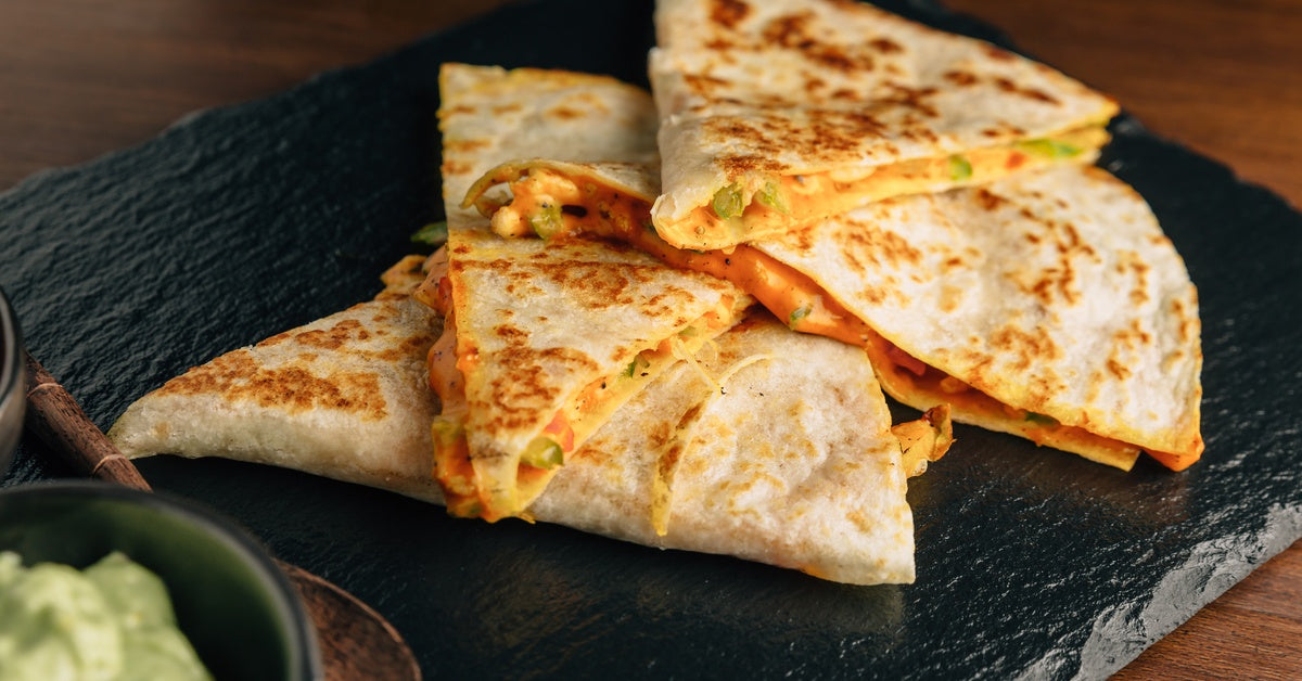 A sliced cheese quesadilla resting on a stone plate next to a small spoon, served with fresh salsa and creamy guacamole.