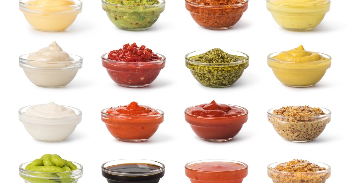 Sixteen small clear bowls stand filled with various sauces and spices of different textures and flavors.