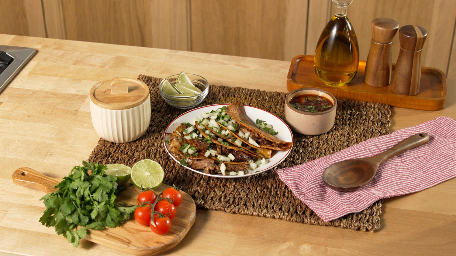 Rise and Puff birria quesadillas served with consommé, lime wedges, cilantro, and fresh tomatoes on a kitchen counter.