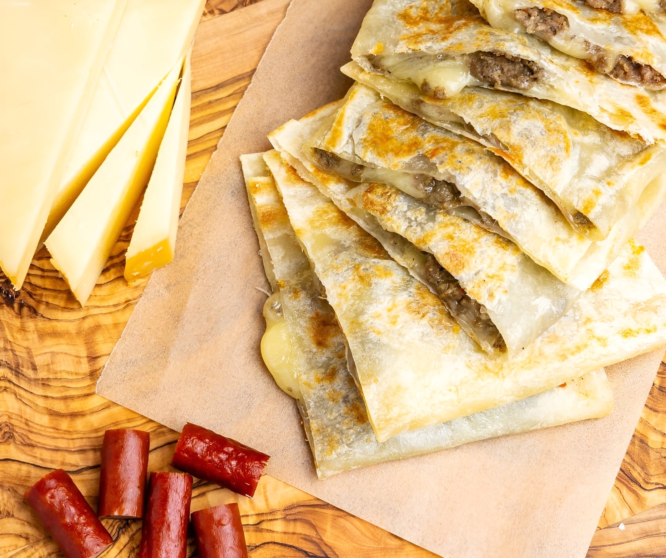 Stacked Rise and Puff quesadilla wedges with melted cheese and meat on parchment, and sausage pieces beside.
