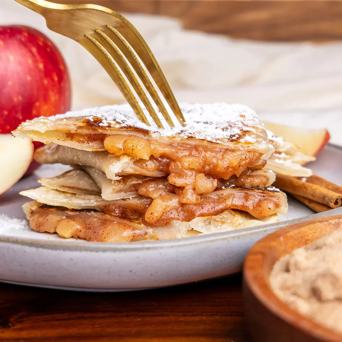 A gold fork pierces a stack of Rise & Puff pies filled with baked spiced apples and dusted with powdered sugar.