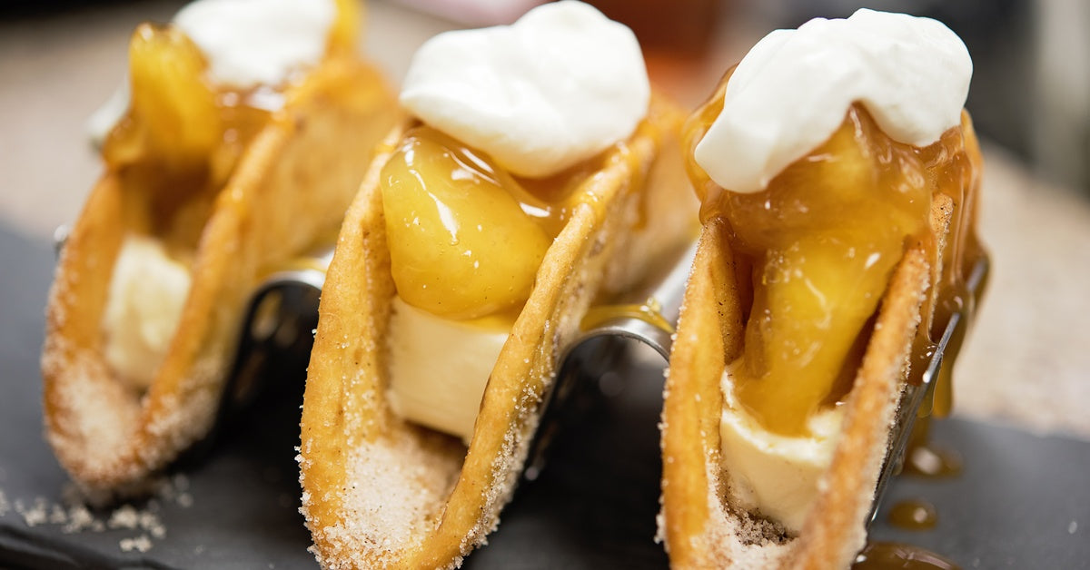 A row of three standing tacos filled with caramelized apples and cream cheese and topped with whipped cream.
