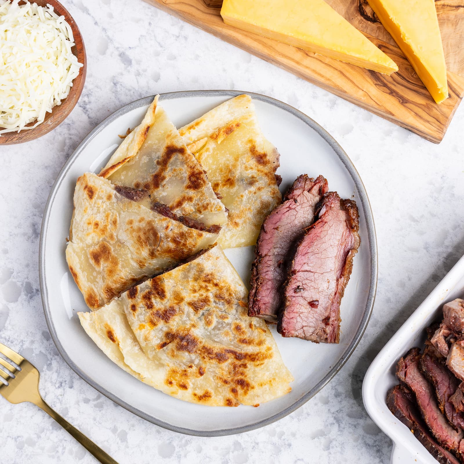 Plate of quesadillas with sliced beef brisket on a light surface with cheese and bread in the background.