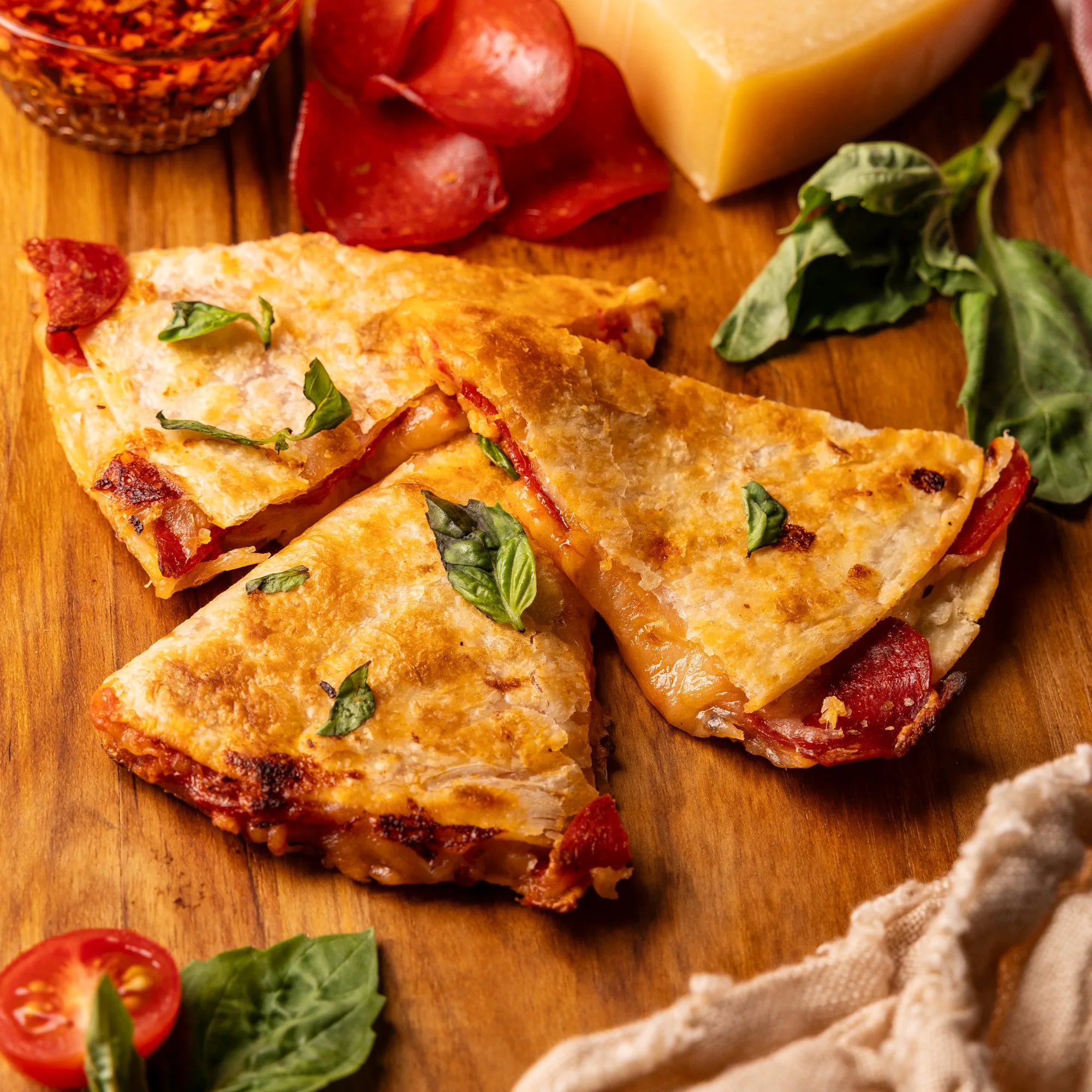 Rise & Puff Uncured Pepperoni Pizza Quesadilla cut into wedges on a wooden board, melted mozzarella and pepperoni showing, styled with fresh basil, Parmesan and chili oil.
