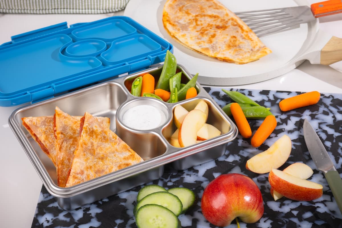 Children's lunchbox with quesadilla pizza slices, fruit, and vegetables on a table.