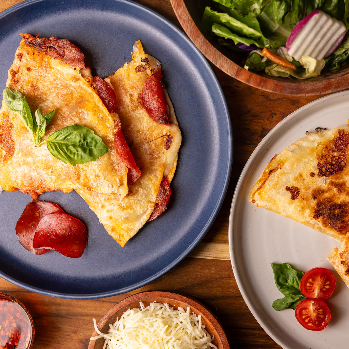 Rise & Puff pizza quesadillas served together: Uncured Pepperoni on a blue plate and Margherita on a white plate, with fresh basil, shredded mozzarella and a side salad.
