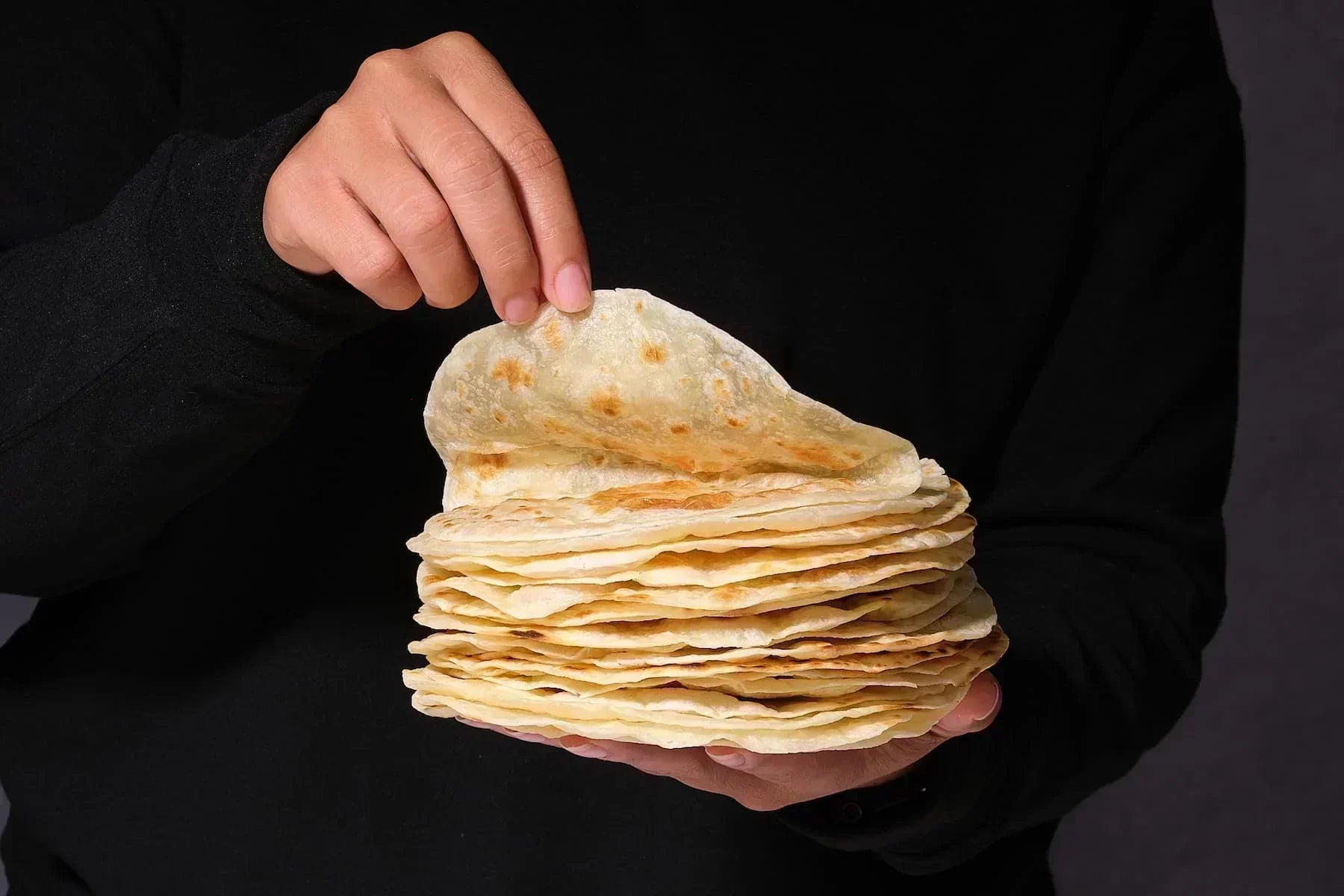 Stack of freshly made tortillas from Rise and Puff held by hand against a dark background.