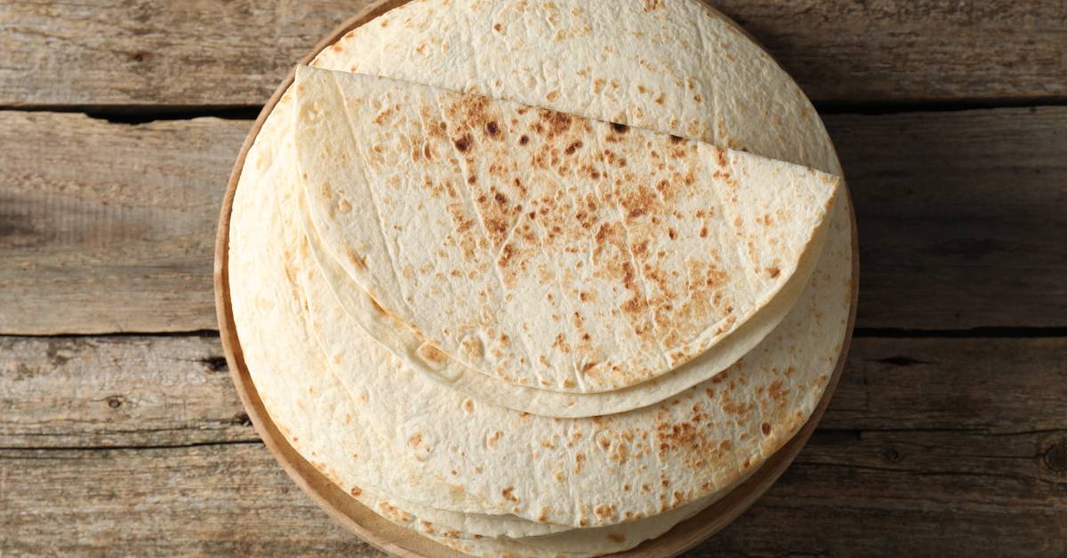 A stack of flour tortillas on a wooden plate. The plate sits on a wooden table and the top tortilla is folded in half.