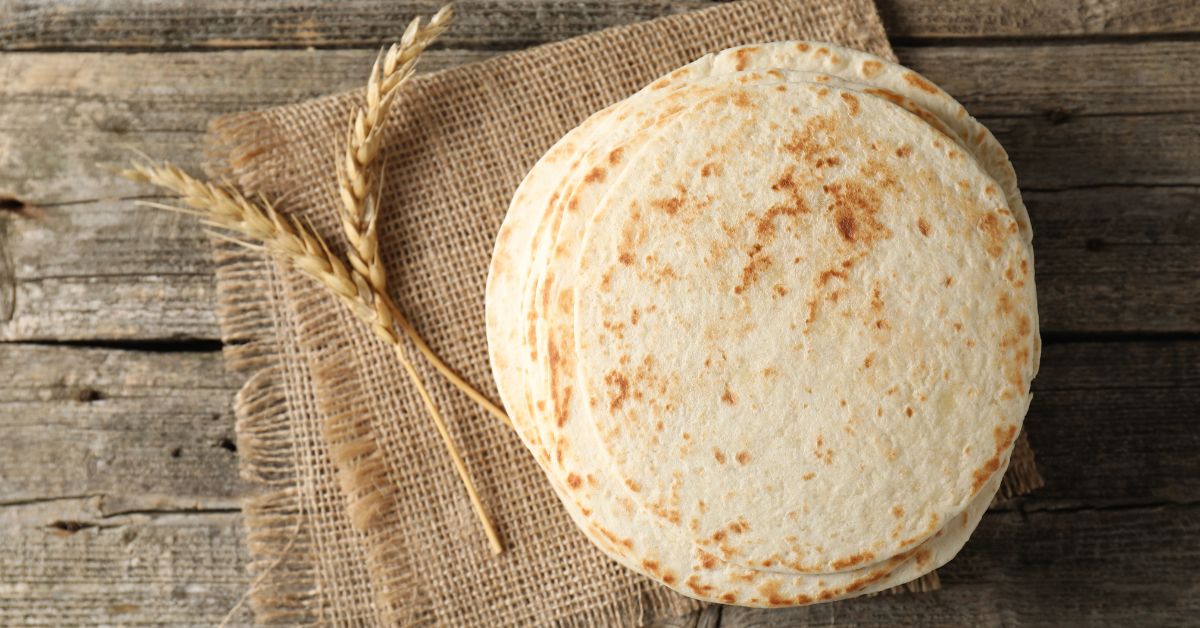 A stack of of flour tortillas lay on two sheets of burlap. Next to the stack, two stems of wheat lay crossed over each other.