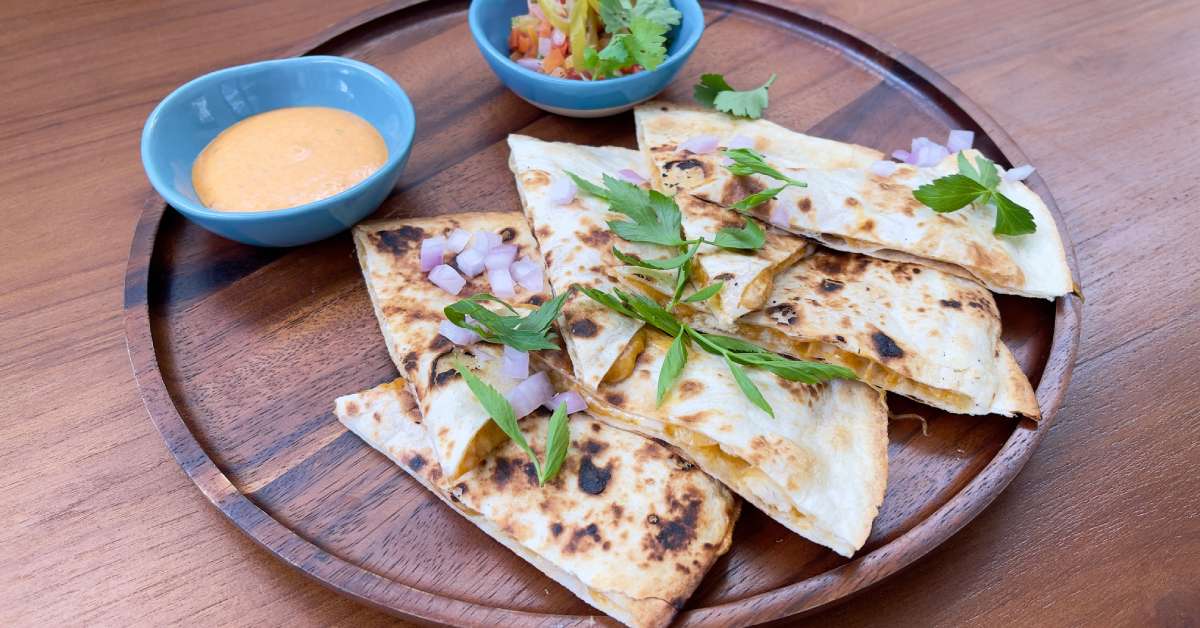 A round wooden plate holds seven pieces of a quesadilla garnished with herbs next to two small bowls of queso and diced peppers.