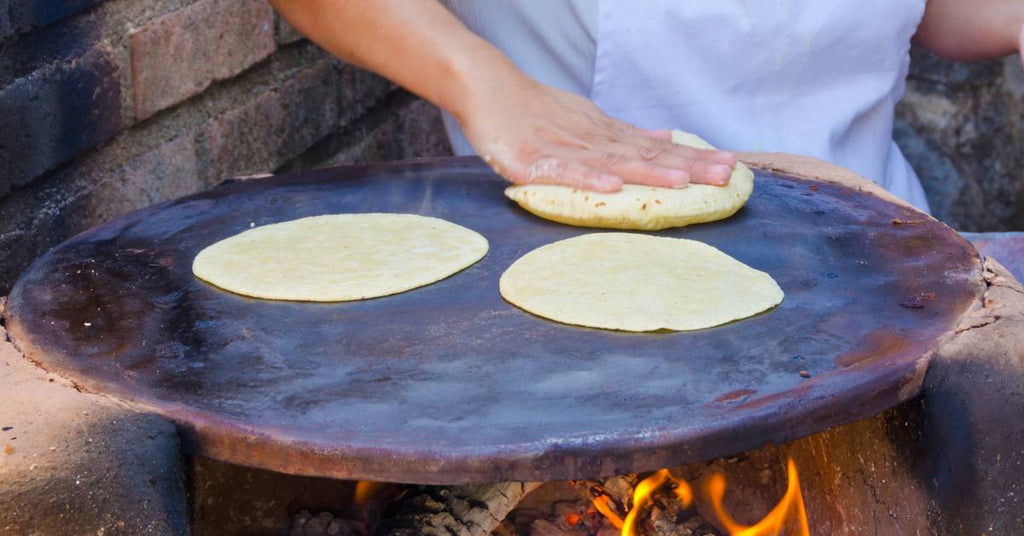 A person using their hands to press one of three tortillas onto a large flat disk with a woodfire burning underneath.