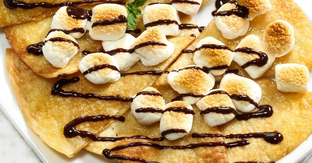 Tortilla slices on a square white plate are topped with toasted marshmallows, a chocolate drizzle, and a green garnish.