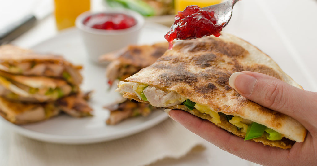 A person holding a portion of a quesadilla containing turkey and peppers while spooning cranberry sauce over the top.