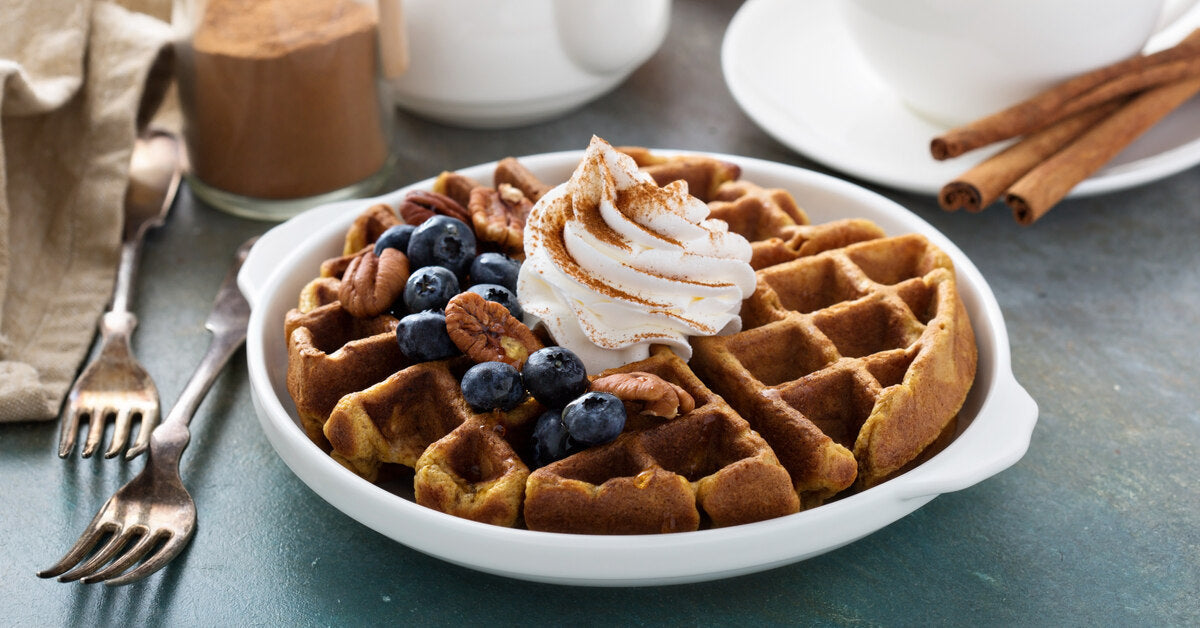 A waffle on a dish is topped with cinnamon-dusted whipped cream, blueberries, and nuts. A mug is next to the dish.