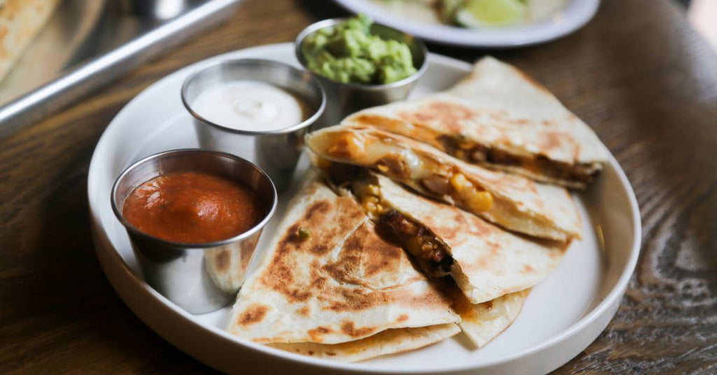 Four quesadilla slices overlap in a row on a deep white plate. Metal dipping cups hold green, white, and red sauces.