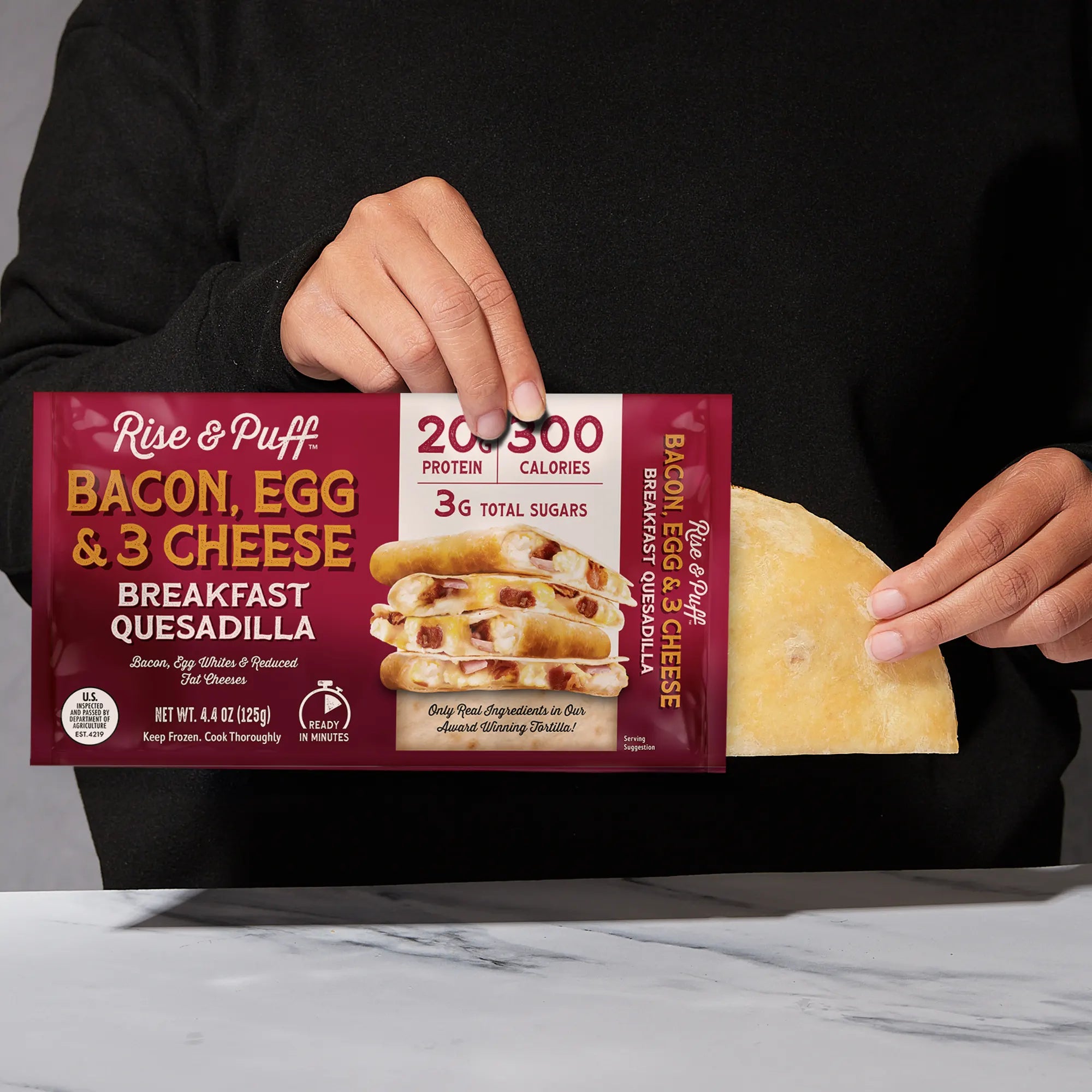 Person holding a Rise & Puff Bacon, Egg & 3 Cheese Breakfast Quesadilla package.