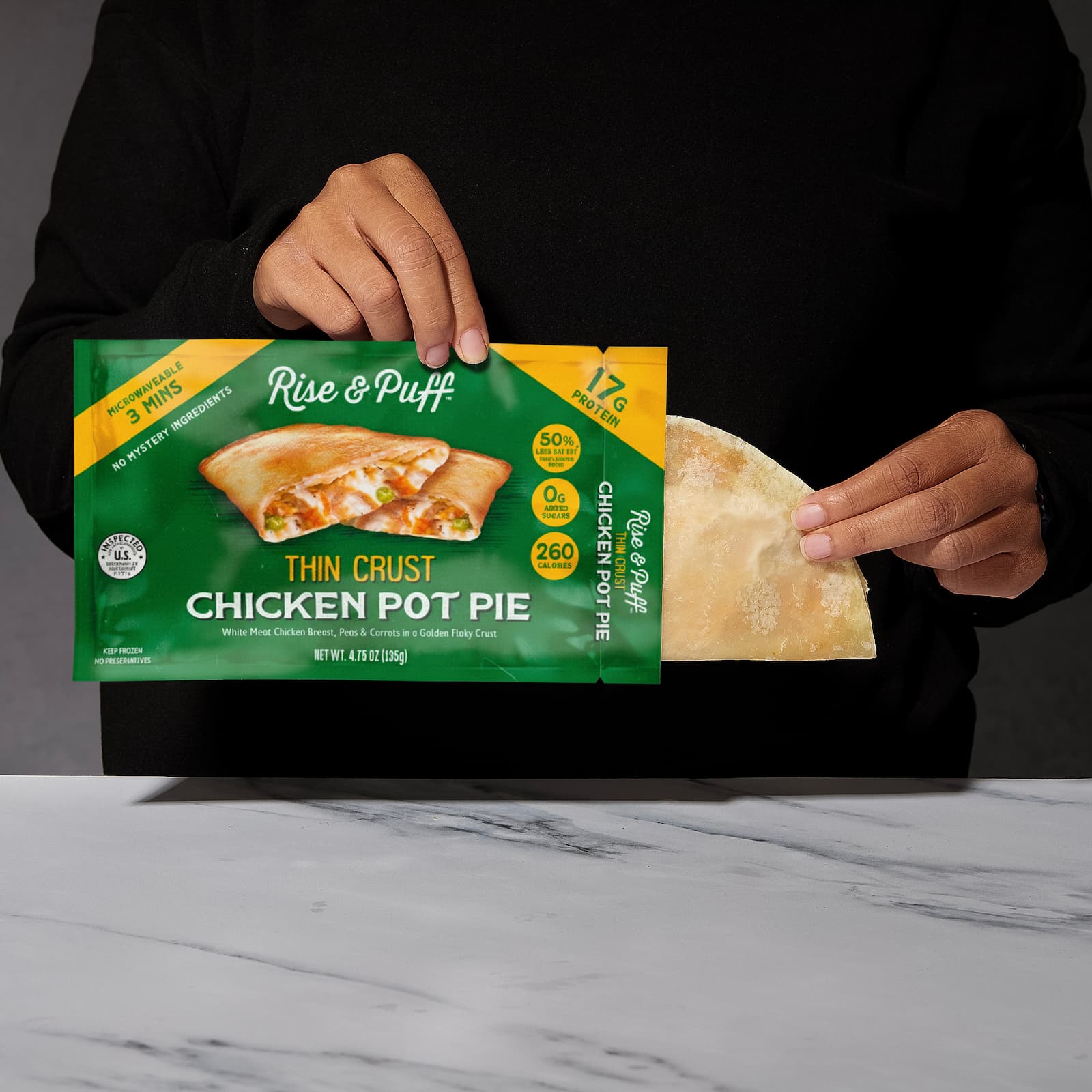 Person holding a Rise & Puff thin crust chicken pot pie package on a marble surface.
