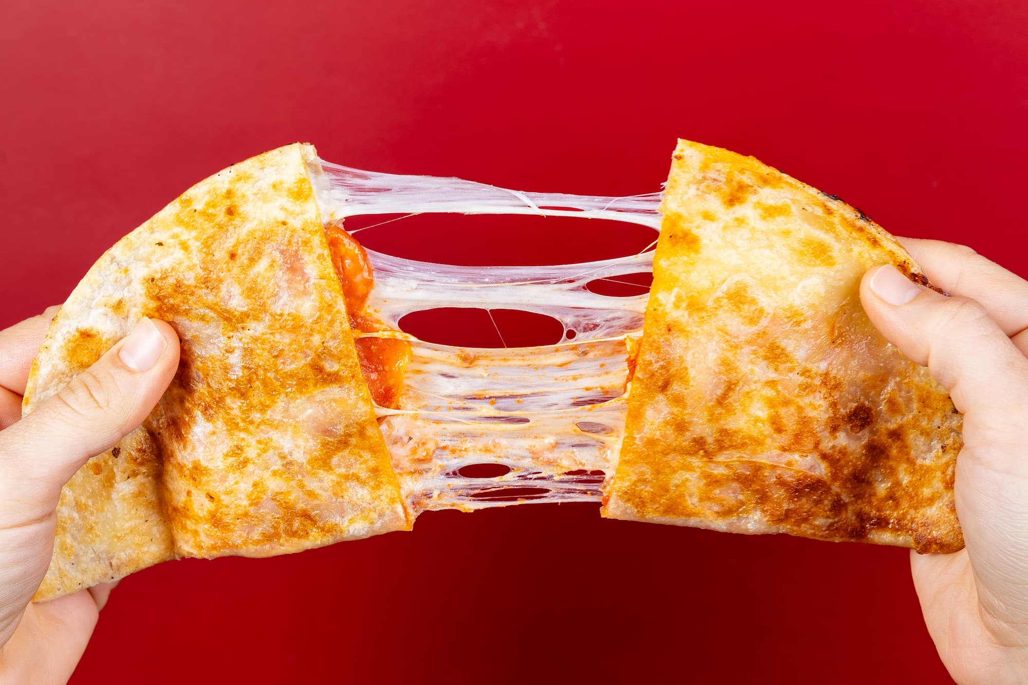 Two hands pulling apart a slice of pepporini pizza quesadilla with stretchy cheese against a red background